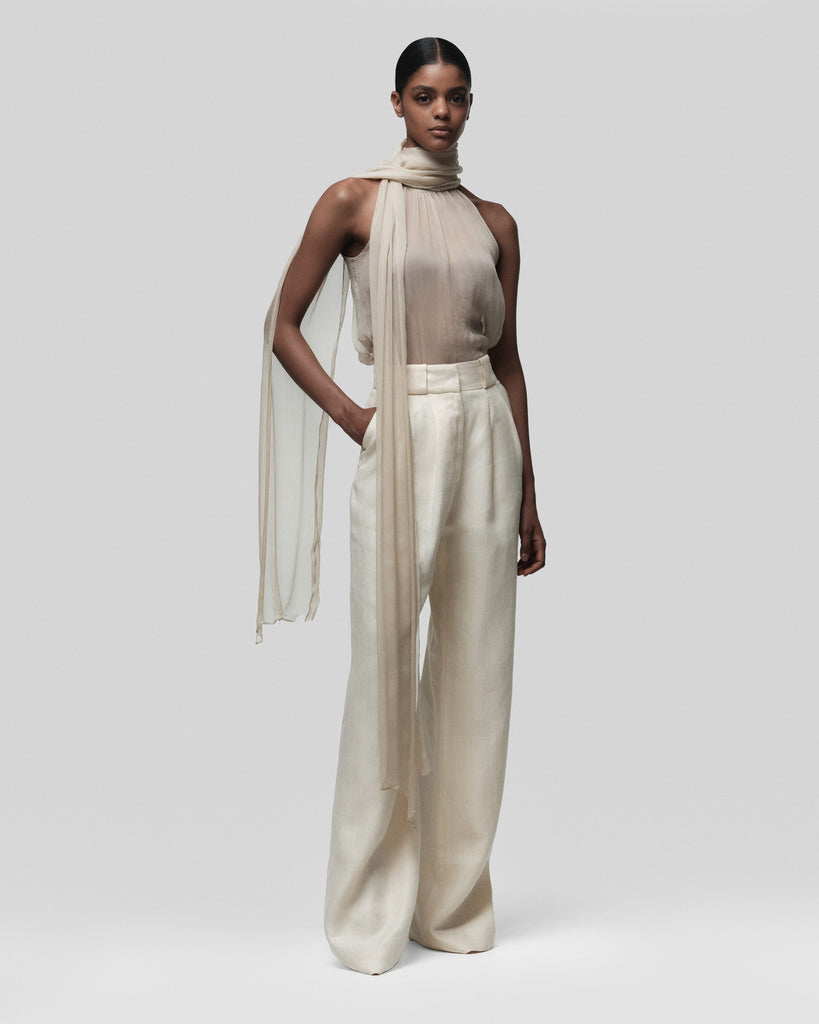 Woman wearing a beige halter top and wide-leg pants with a sheer scarf on a white background