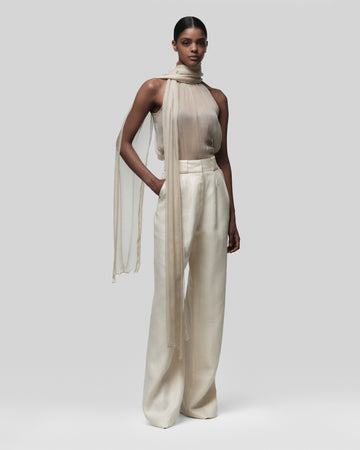 Woman wearing a beige halter top and wide-leg pants with a sheer scarf on a white background