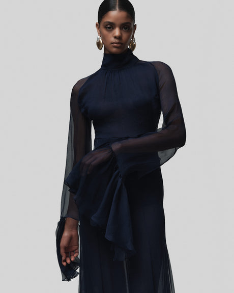 Woman wearing a dark blue dress with sheer sleeves against a gray background